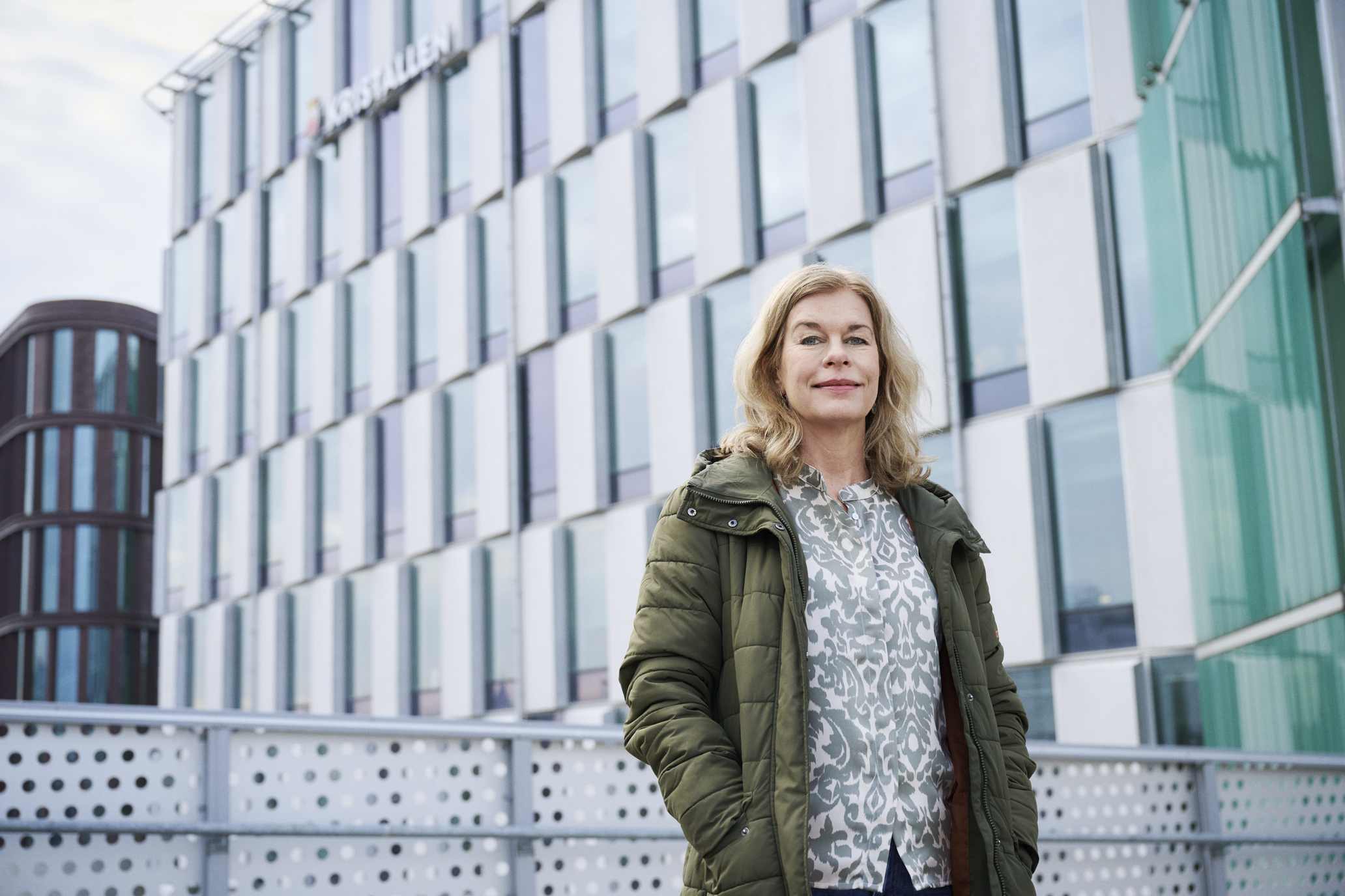 A blonde woman stands with her hands in her jacket pockets in front of a glass building