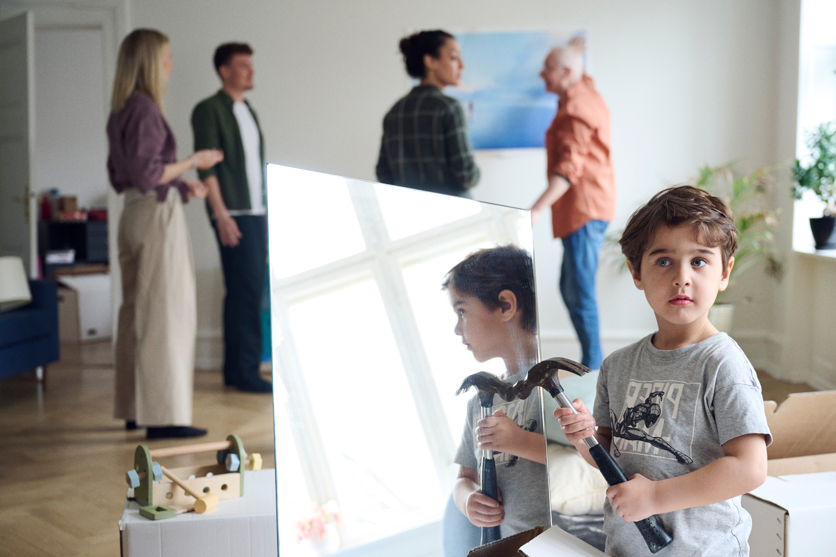 A young child stands in front of a mirror holding a hammer while four adults speak in the background