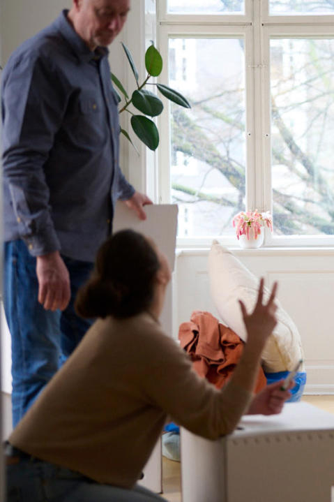 A child in the background knocks over a vase of flowers while two adults speak in the foreground